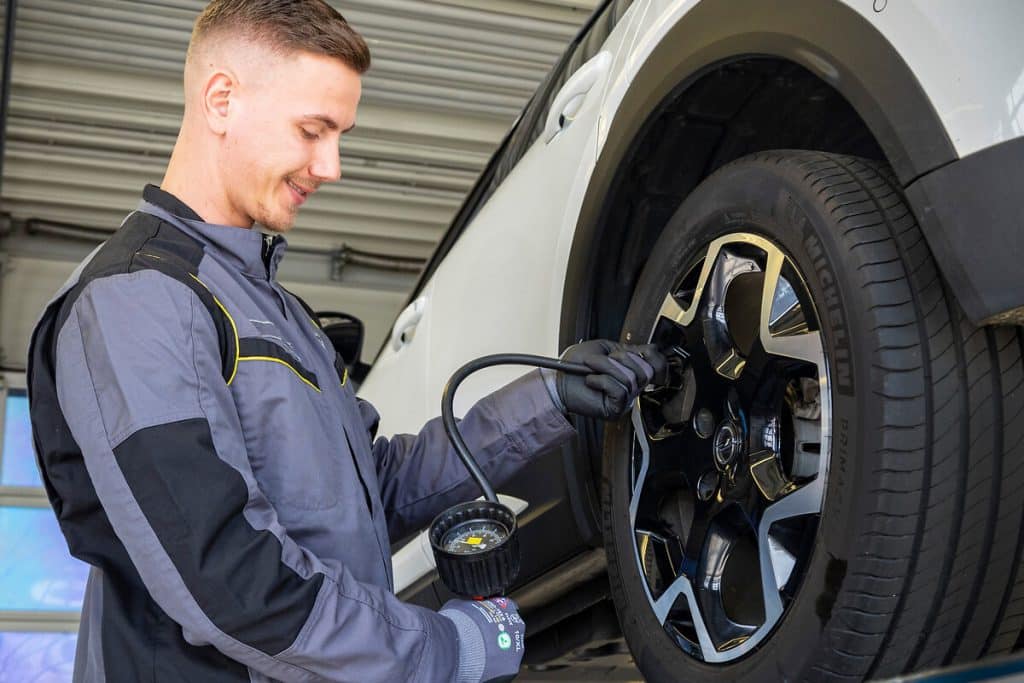 Ein Mann mit grauer Arbeitskleidung schraubt an der Felge eines weißen Auto der Marke Opel